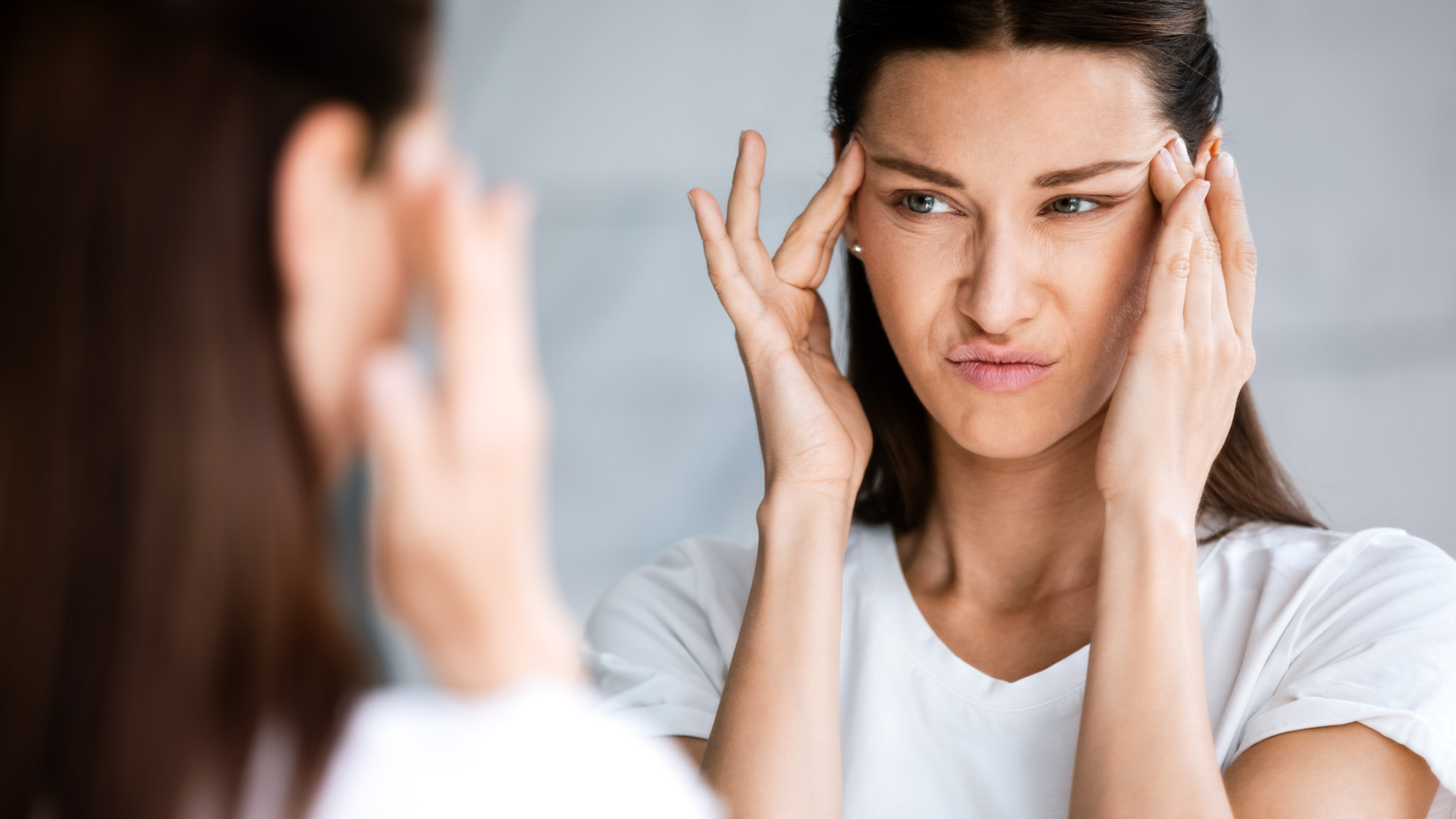 Woman looking into a mirror with her fingers on her temples because of the negative thoughts in her mind.